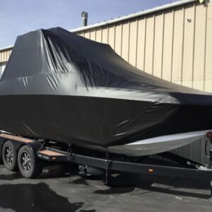 A boat covered with a black protective tarp is parked on a trailer outside a metal building.