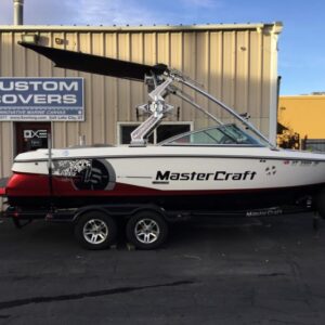 A MasterCraft speedboat on a trailer, parked in front of a building with a sign reading "Custom Covers." The boat has a canopy and a detailed graphic design on the side.