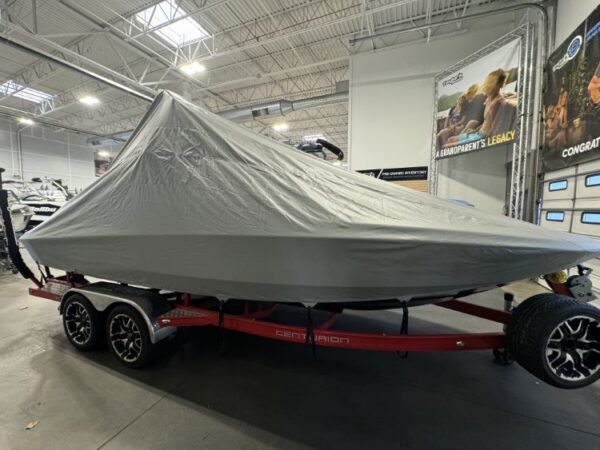 A boat on a red trailer is covered with a gray tarp in a large indoor space with bright lighting.