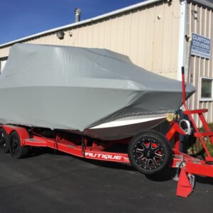 A boat with a gray cover sits on a red trailer parked outside a building labeled "Custom Covers.