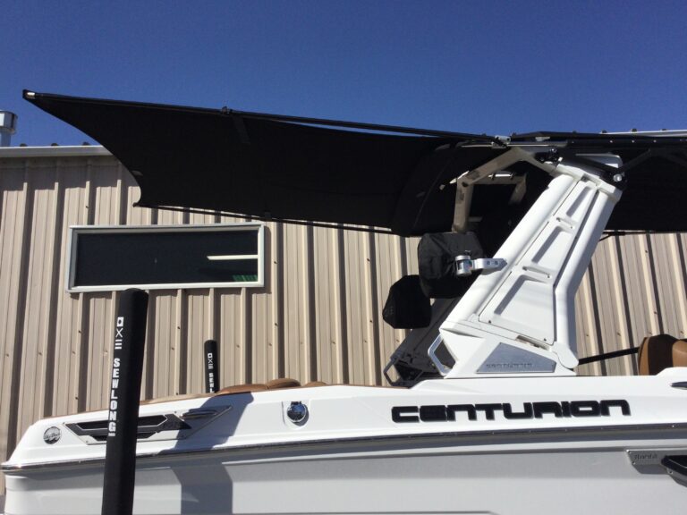 A white Centurion boat is parked near a beige metal building, featuring a raised black canopy, wakeboard tower, and an adjustable SewLong shade sail for enhanced comfort.