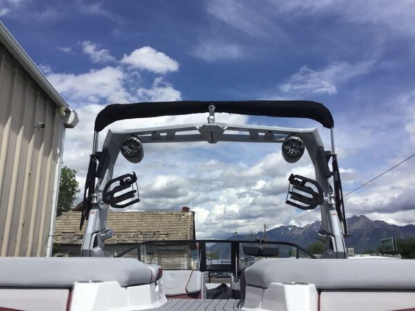 View from the rear of a boat with a wakeboard tower, speakers, and board racks, against a cloudy sky and mountain backdrop.