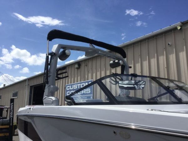 A white boat with a wakeboard tower is parked in front of a metal building under a blue sky with clouds.