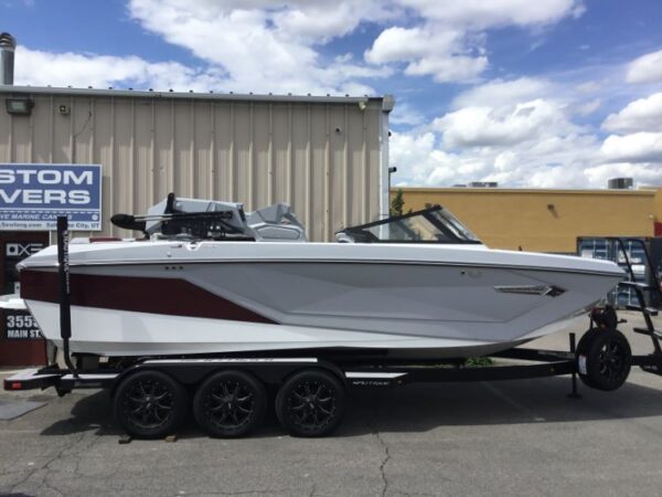 A white and maroon speedboat on a black trailer is parked outside a metal building under a partly cloudy sky.