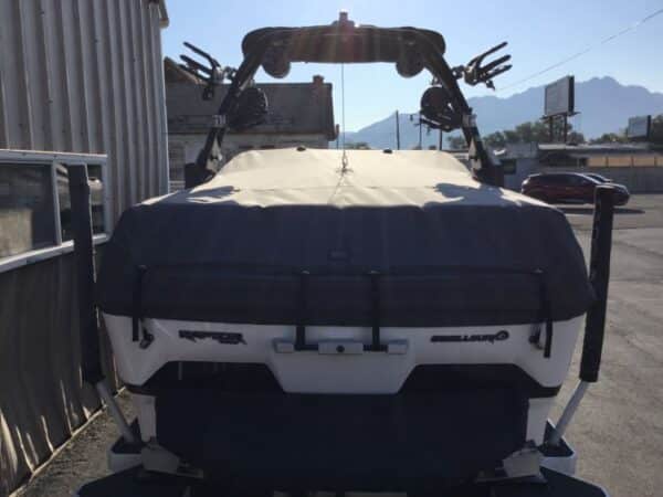 Back view of a covered boat on a trailer, parked near a building. Mountains are visible in the background.