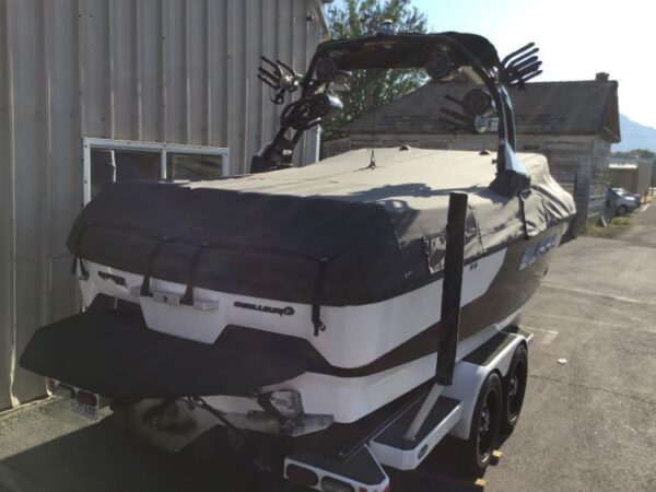 A white and black boat on a trailer is parked outside a metal building. The boat is covered and has speakers mounted on a black frame.