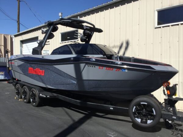 A black and gray Malibu wakeboard boat with red accents is parked on a black trailer in front of a beige industrial building.