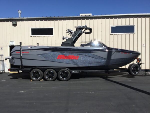 A gray and black motorboat with the brand name "Malibu" in red lettering is parked on a multi-axle trailer in front of a beige industrial building.