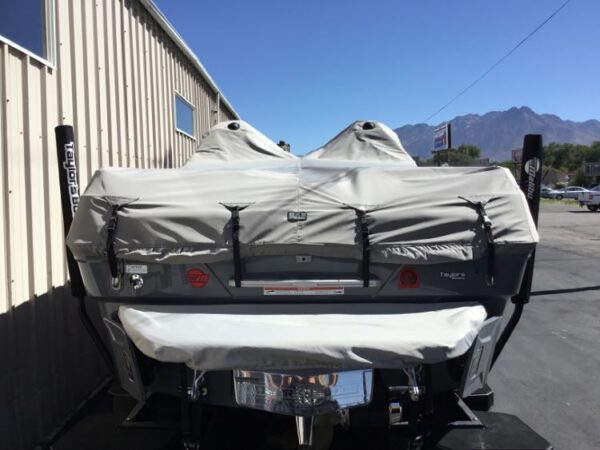 A covered boat is parked beside a metal building on a sunny day. The boat has gray protective covers over its engines and a white cover over the main body. Mountain range visible in the background.