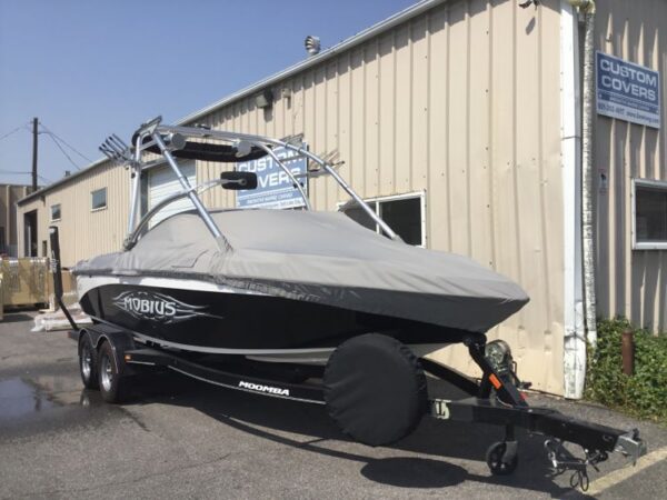 A black Moomba Mobius boat is on a trailer outside a beige building with "Custom Covers" signs. The boat is covered with a gray protective cover.