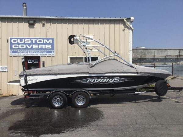 A black and white Mobius LSV boat is covered and parked on a trailer outside a building with a "Custom Covers" sign.