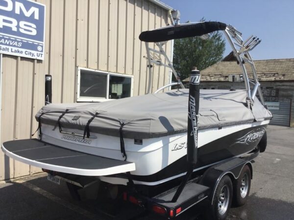 A wakeboard boat with a gray cover is parked on a trailer outside a building. A sign reads "Salt Lake City, UT.