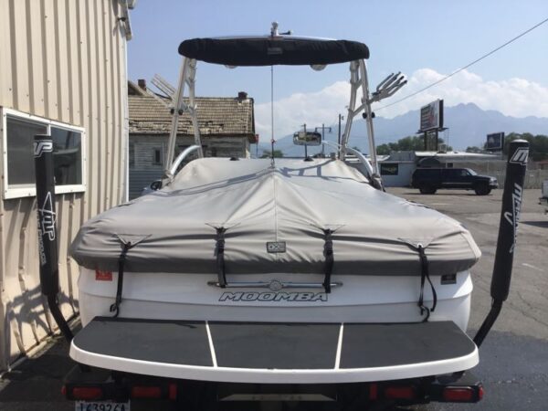 A boat covered with a gray tarp is parked beside a building. Two wakeboard racks and a tower are visible. Mountains and a vehicle appear in the background.